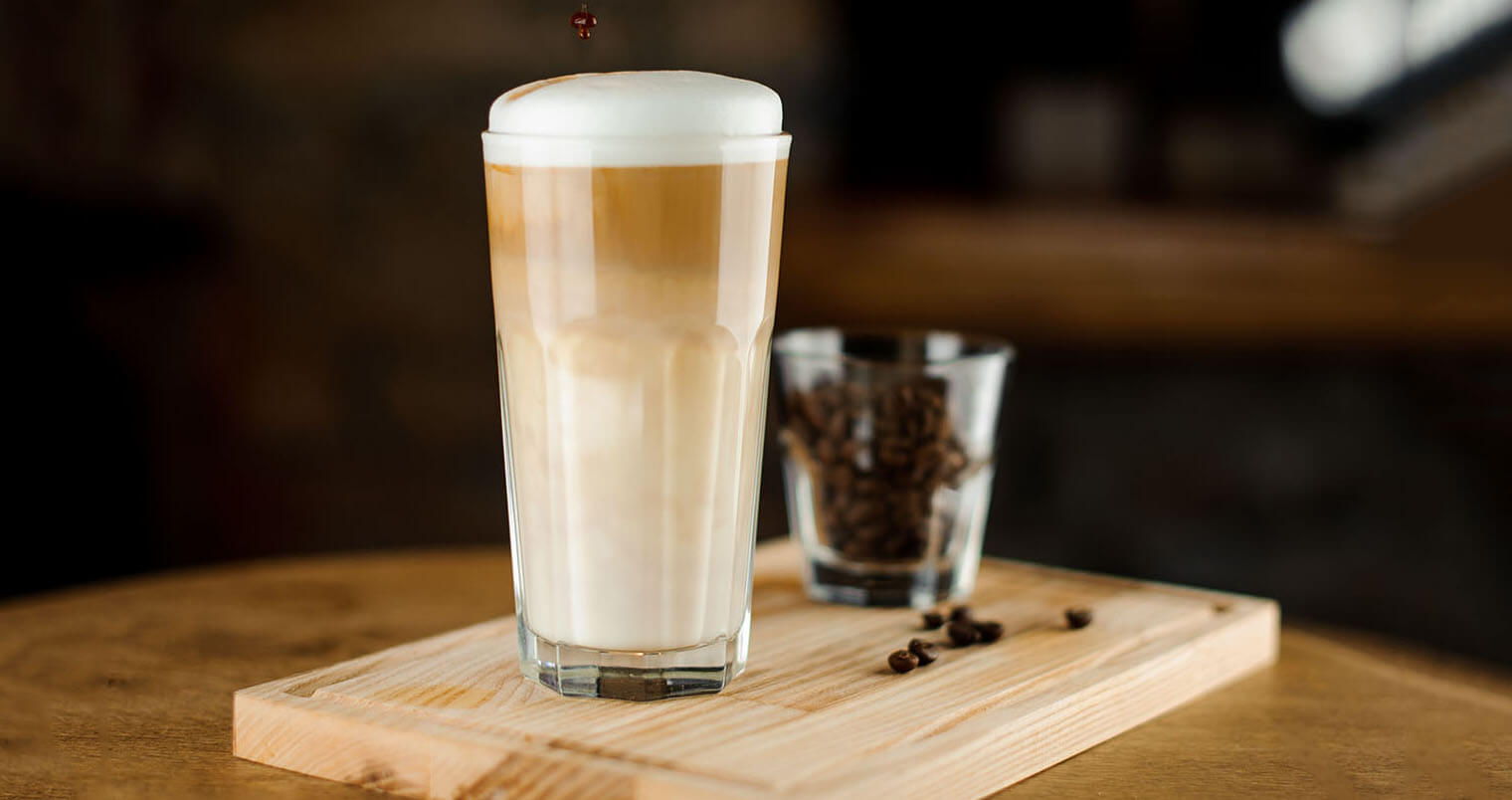 Hot coffee latte in a high glass glass on a wooden board. Glass of coffee beans in the background