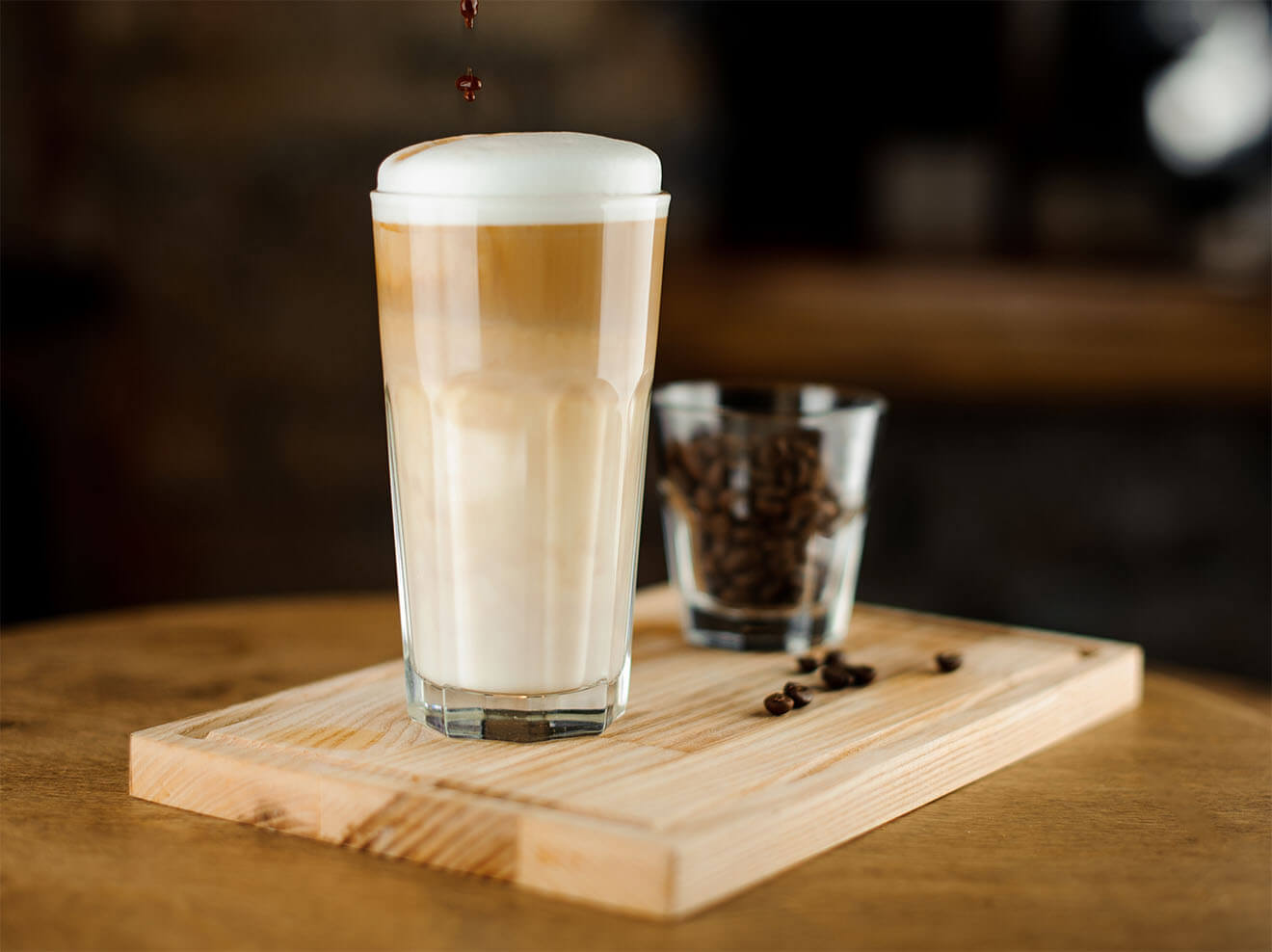 Hot coffee latte in a high glass glass on a wooden board. Glass of coffee beans in the background