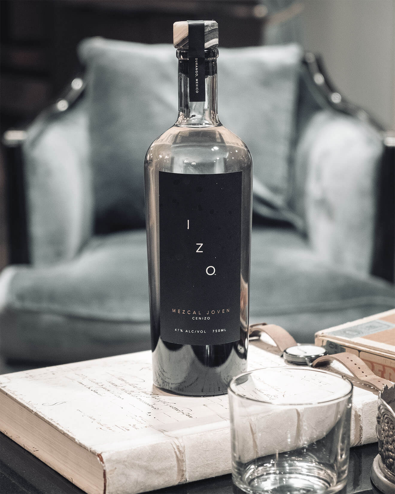 IZO Mezcal Joven, bottle on book, glasses, grey chair