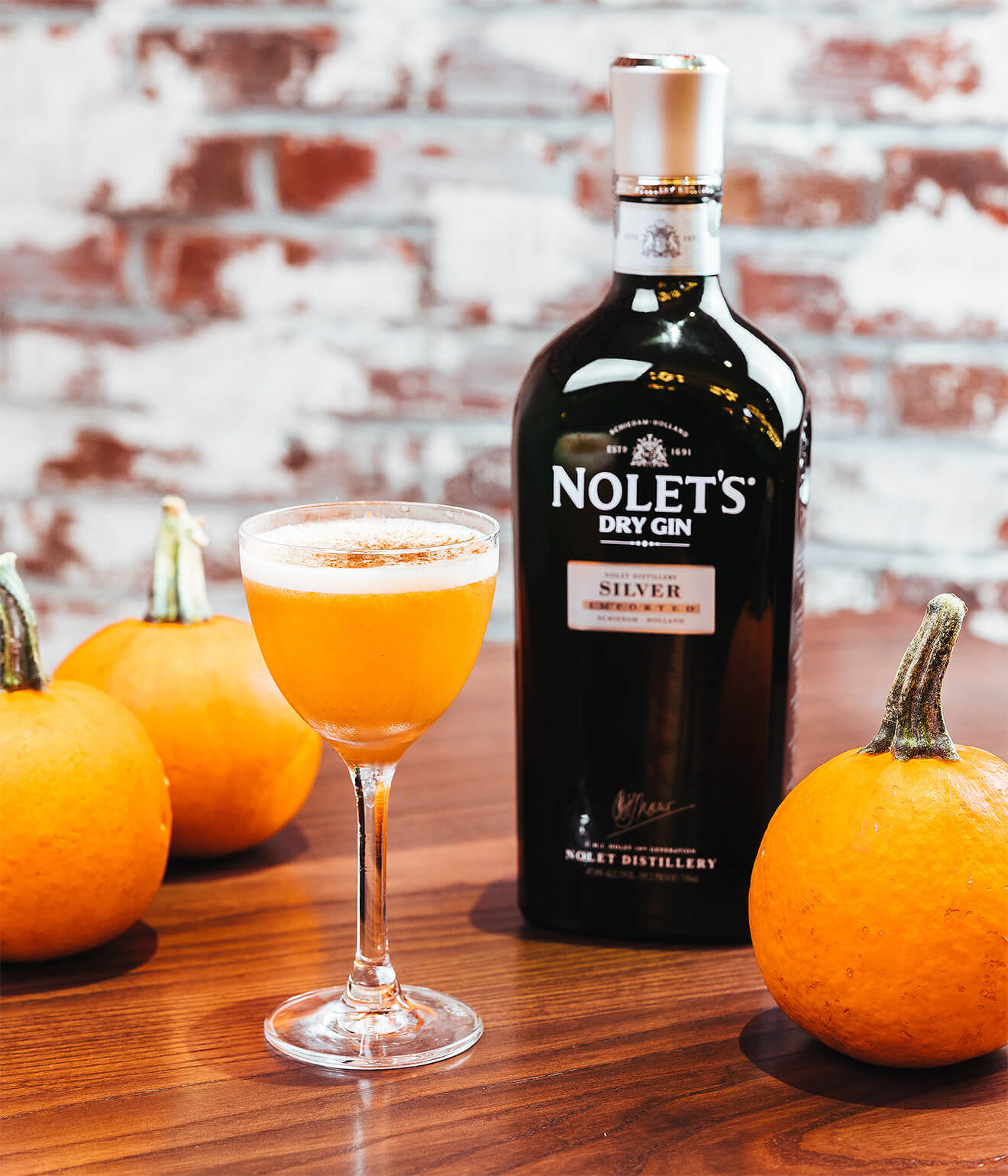 NOLET’S Silver Pumpkin Flip, cocktail, bottle and pumpkin garnish, brick wall background