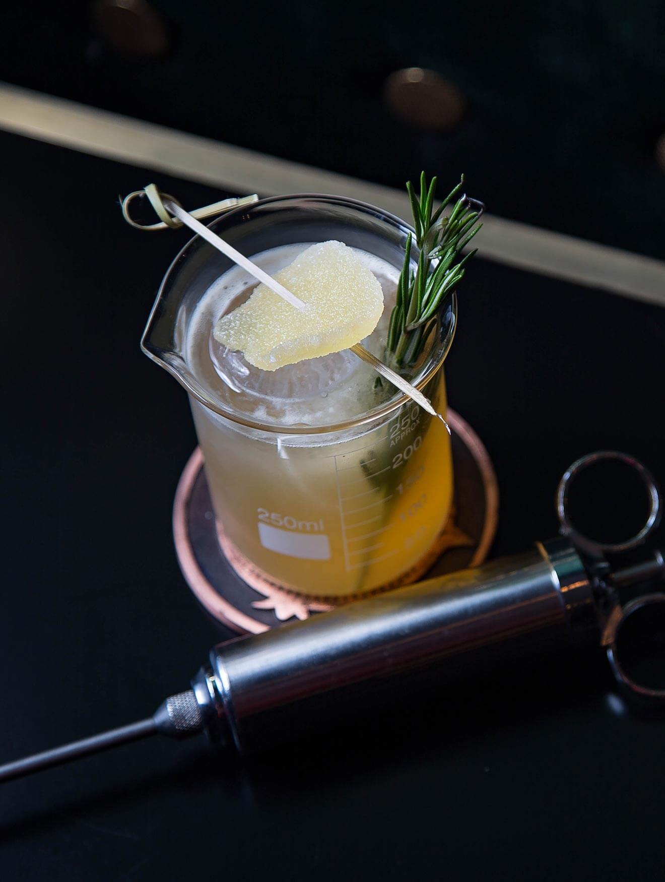 The Peaty Penicillin Cocktail, dark back with syringe and garnish