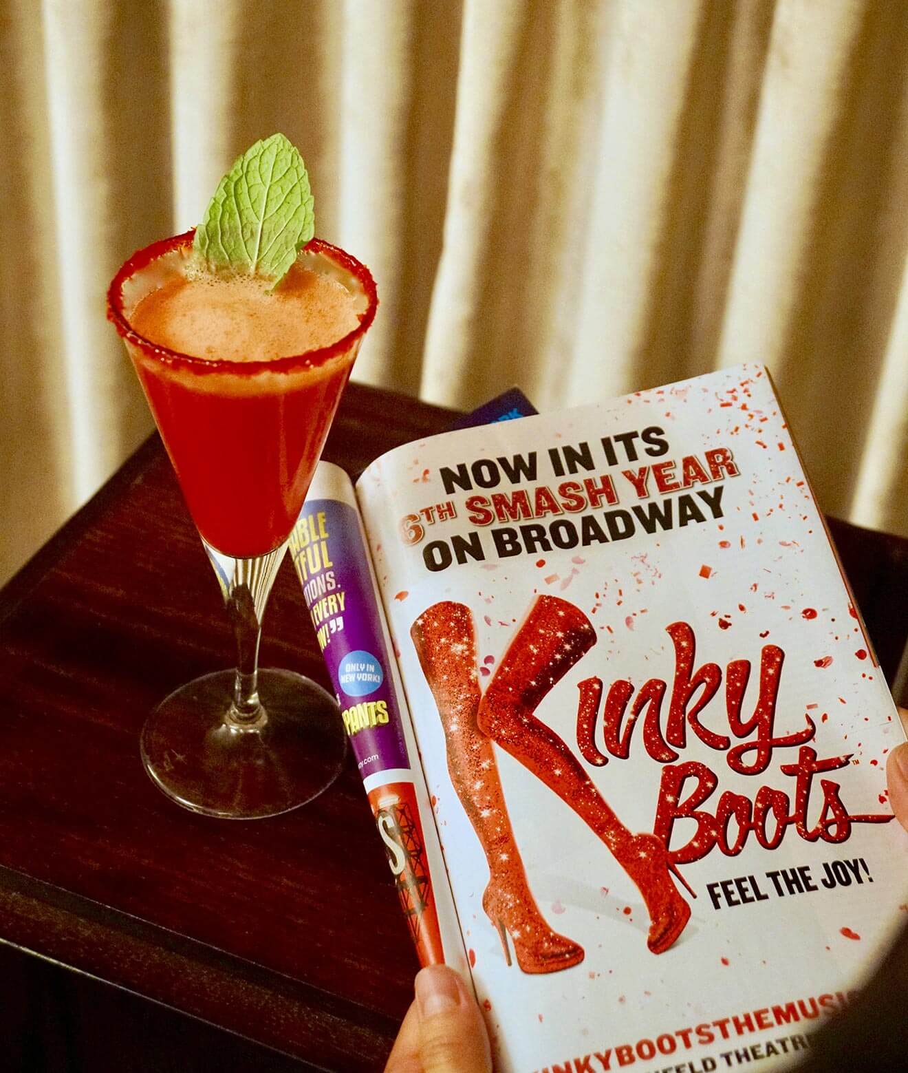 The Shaken Stiletto, Kinky Boots, cocktail and book on table with curtain in back